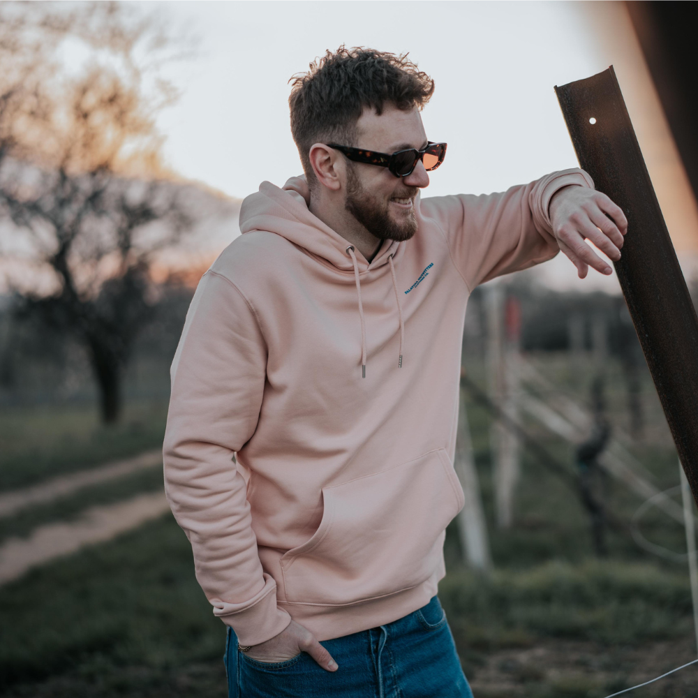 Ein Model steht lachend angelehnt an einer Eisenstange in den Weinbergen und trägt dabei einen rosafarbenen Hoodie und eine VINO VISTA Sonnenbrille Chardonnay