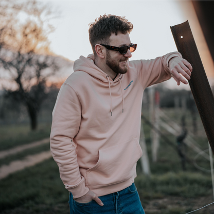 Ein Model steht lachend angelehnt an einer Eisenstange in den Weinbergen und trägt dabei einen rosafarbenen Hoodie und eine VINO VISTA Sonnenbrille Chardonnay