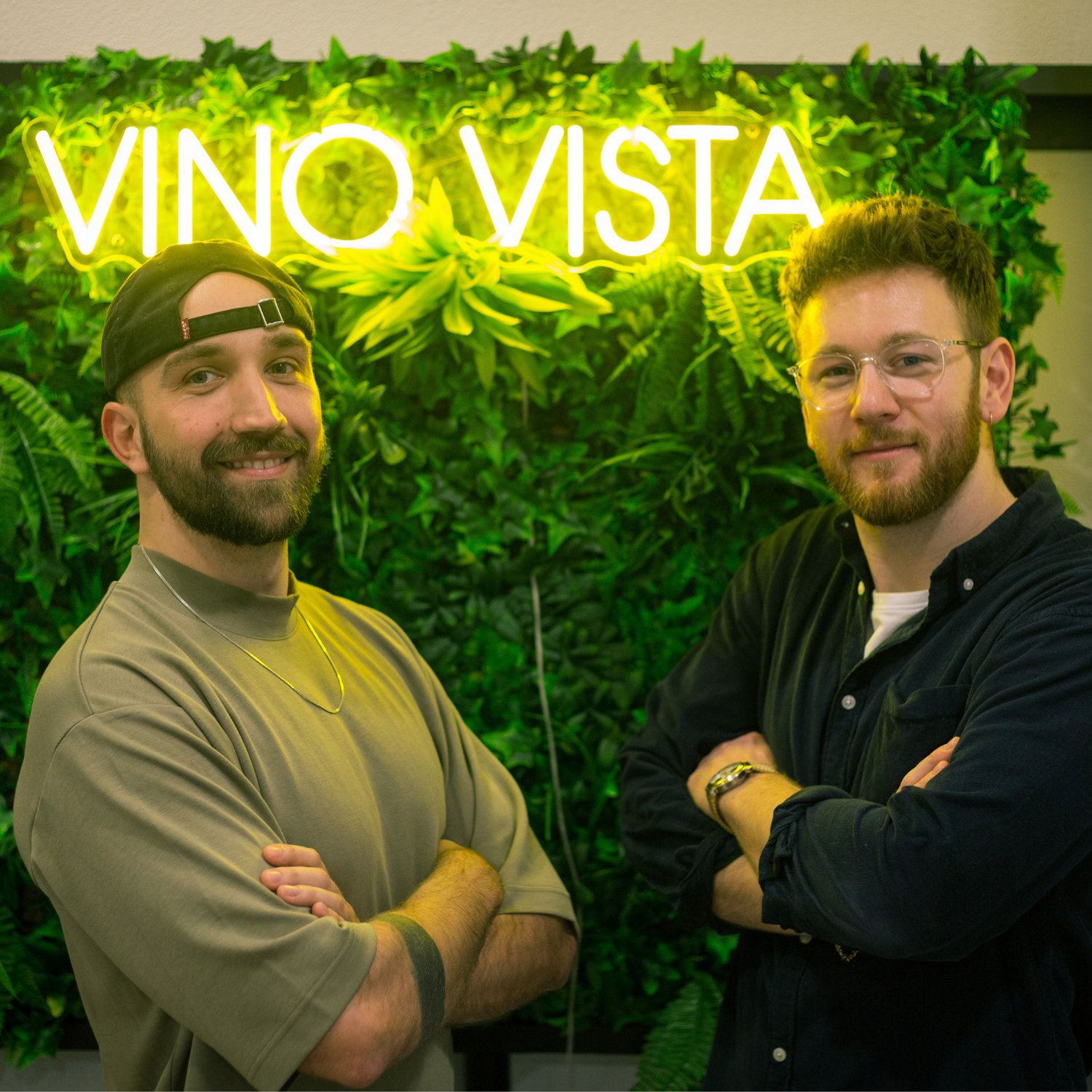 Gründer Leander und Moritz stehen nebeneinander mit armen verschränkt. Im Hintergrund leuchtet das VINO VISTA Logo